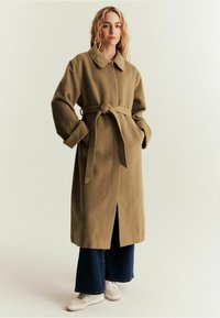 Olivgrüner Mantel aus Wollmischung mit Kragen, langen Ärmeln und Taillengürtel. Verfügt über Seitentaschen und einen vorderen Schlitz. Kombiniert mit jeans mit weitem Bein und Turnschuhen.