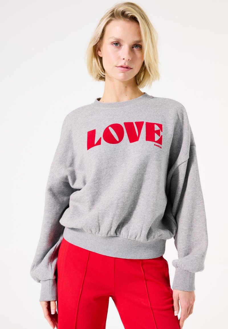 Sudadera gris con gráfico rojo de "LOVE", presenta un cuello redondo, corte relajado y cintura fruncida. Combinada con pantalones rojos.