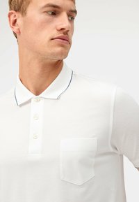 Witte polo met een kraag, drieknoopsluiting, marineblauw accent en een borstzak. De stof is textured en lichtgewicht.