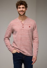 Chemise henley à manches longues, rayée rouge, avec patte de boutonnage à trois boutons et tissu texturé. Présentant un petit logo et une coupe décontractée. Portée avec un jean foncé.