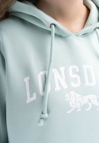 Šviesiai mėtinės žalios spalvos džemperis su užtrauktuku, priekyje vaizduojantis užrašą „LONSDALE“ baltos spalvos, su kapišonu ir sidabriniais aklūnais. Minkšta tekstūra.