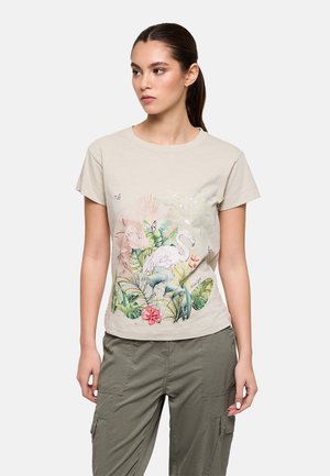 Mujer con cabello oscuro recogido en una coleta, que lleva una camiseta beige de manga corta con un estampado colorido de flamencos y plantas tropicales, y pantalones cargo oliva.
