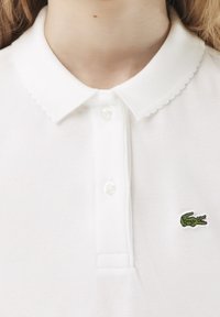 Valkoinen polo-paita, joka on valmistettu tekstuurista kankaasta. Paidassa on aaltoileva kaulus ja kaksi nappia sekä pieni vihreä kirjailtu krokotiililogo.