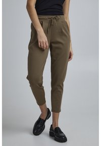 Khaki joggingové kalhoty s páskem na stažení, zúženými nohavicemi a bočními kapsami. Nosené s černými mokasíny na neutrálním pozadí.