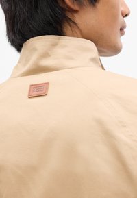 Μπεζ μπουφάν με ψηλό γιακά και δερμάτινη ετικέτα στο πίσω μέρος, με λεία, ματ υφή και καθαρές λεπτομέρειες ραφής.