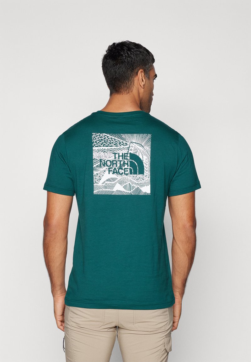 Teal bomull t-skjorte med et kvadratisk grafikk på baksiden som viser teksten "The North Face" og abstrakte linjemønstre i hvitt. Korte ermer, rund hals.