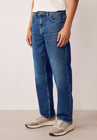 Blugi din denim albastru cu un croi drept, având un design clasic cu cinci buzunare și decolorări subtile. Asortați cu adidași gri și bej.
