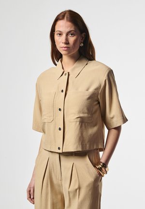 Woman with red hair wearing beige short-sleeve button-up shirt and matching high-waisted pants, accessorized with gold bracelets and earrings.