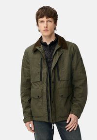 Olivengrøn jakke med brun fløjlskrave, forlommer med knapper foran og et sort inderlag. Har lynlås og trykknaplukning.