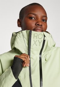 Giacca impermeabile verde chiaro con colletto alto, zip nera, logo Roxy in dettaglio perforato, e polsini con foro per il pollice in contrasto nero.