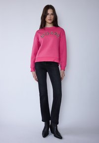 Rosa Sweatshirt mit dem Aufdruck "MILANO" vorne, kombiniert mit schwarzen Jeans und spitzen schwarzen Stiefeletten. Glatte Textur, lässige Passform.