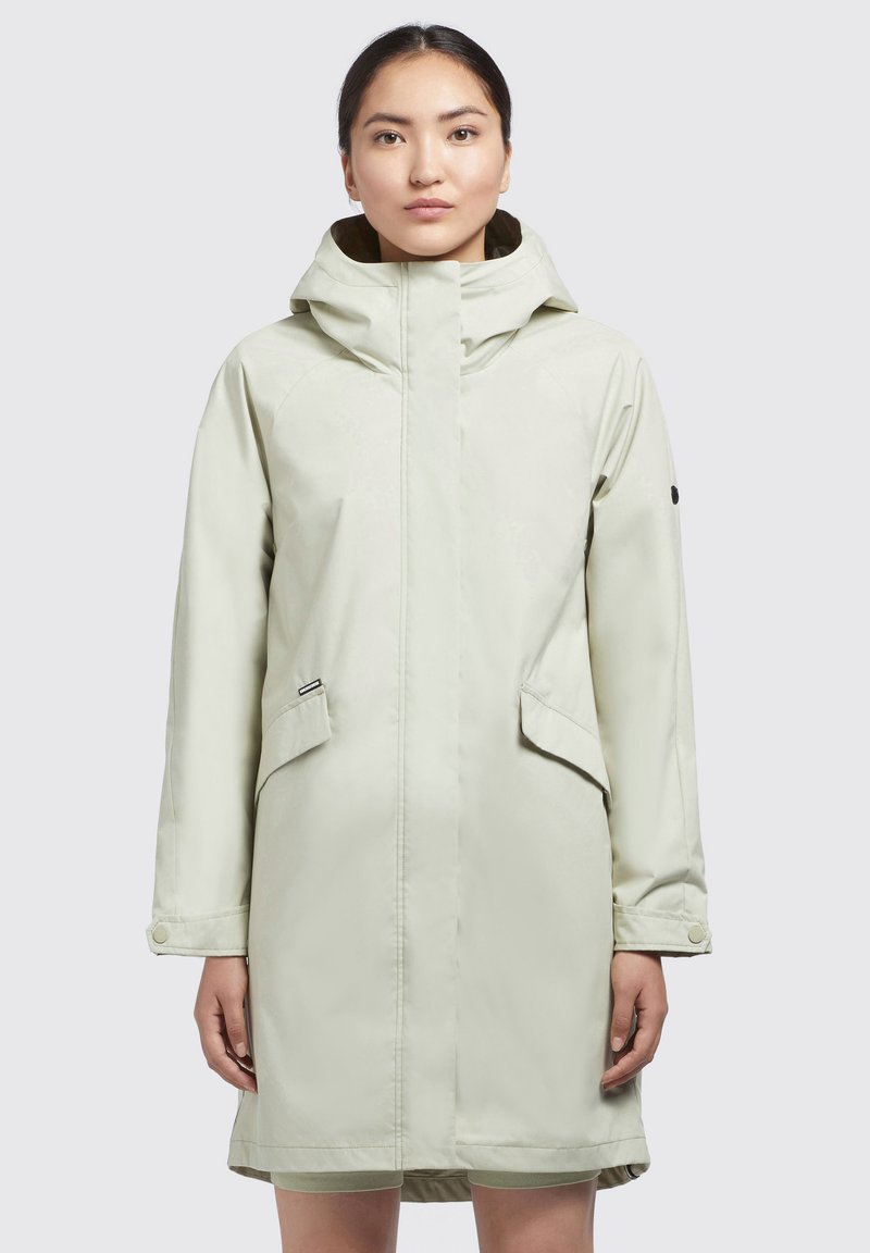 Parka impermeabil de culoare verde deschis, cu glugă, buzunare laterale și manșete cu capse. Prezintă o textură netedă și un design minimalist. Fundal simplu.