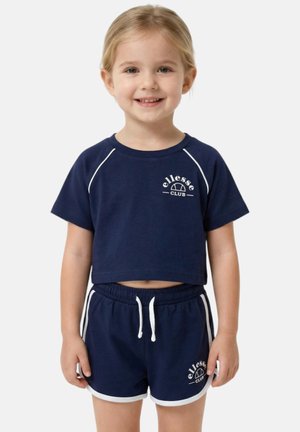 Jeune fille souriante aux cheveux blonds portant un t-shirt court ellesse Club bleu marine et un short assorti avec un liseré blanc sur fond uni.