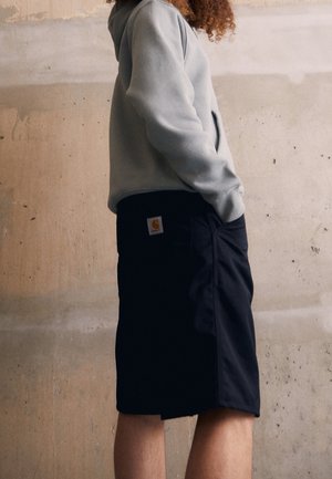 Shorts noirs avec poches, en tissu robuste, arborant un patch logo visible. Associés à un sweat à capuche gris clair.