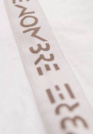 Witte stof met een gestructureerde beige streep waarop stempels in bruin letters en symbolen zijn aangebracht, diagonaal over het materiaal geplaatst.