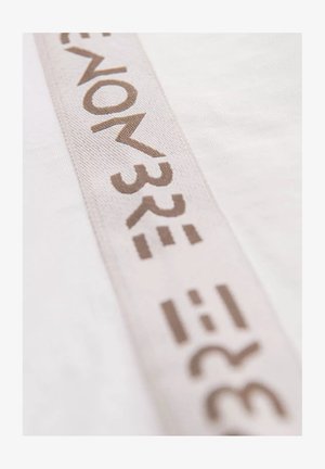 Tissu blanc avec une bande beige texturée présentant des lettres et des symboles bruns estampés, positionnés en diagonale sur le matériau.