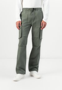 Person wearing olive green cargo pants with drawstring waist and white sneakers, standing against a white background.