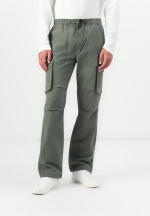 Person wearing olive green cargo pants with drawstring waist and white sneakers, standing against a white background.