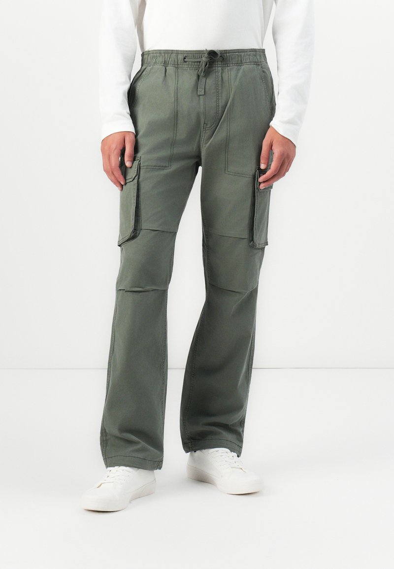 Person wearing olive green cargo pants with drawstring waist and white sneakers, standing against a white background.