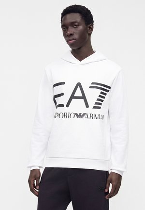 Νεαρός άνδρας που φοράει λευκή φούτερ EA7 Emporio Armani με μαύρο παντελόνι, στέκεται μπροστά σε απλό ανοιχτό φόντο.