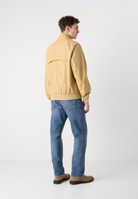 Lys gul bomberjakke med elastiske manchetter og kant, båret over blå jeans og brune sko, set fra ryggen. Simpelt design, glat tekstur.