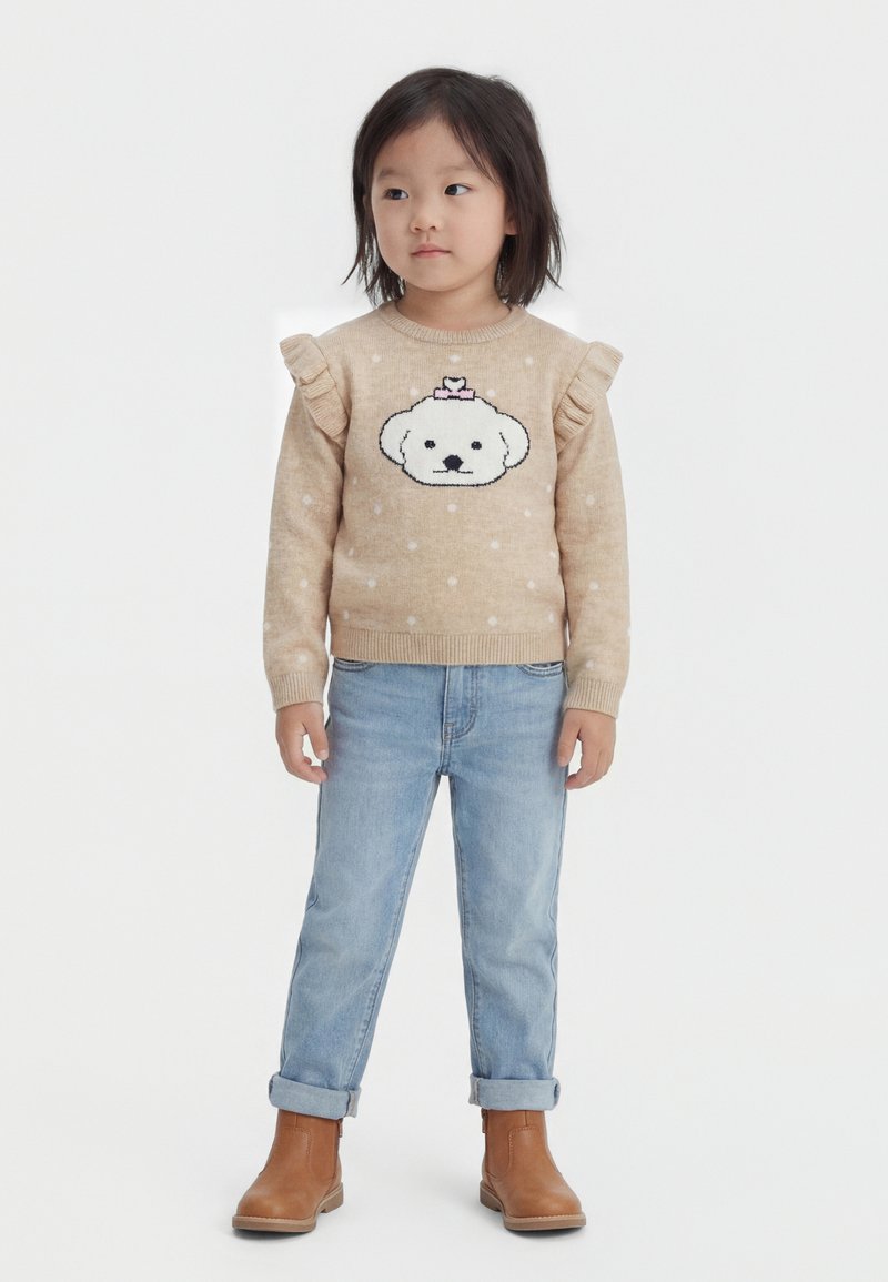 Jeune enfant aux cheveux mi-longs portant un pull beige avec un visage de chien, un jean bleu clair retroussé et des bottes marron, debout sur fond blanc.