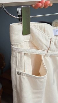 Hand som håller en vit denimbyxa som är klämd på en metallklämmare, vilken visar detaljerna på framlappen och bältesöglorna.