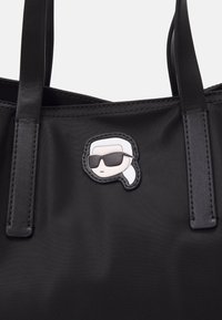 Schwarze Tragetasche aus glattem Nylon, mit einem abnehmbaren Patch einer Figur mit Sonnenbrille. Zwei robuste Lederträger zum Tragen.