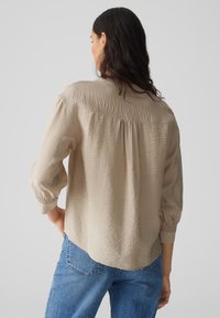 Beige, textured blouse with a loose fit, long puffed sleeves, and a slightly curved hem, featuring a gathered back detail. Worn with blue jeans.