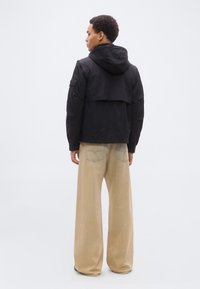 Άνδρας που φοράει μαύρο μπουφάν με κουκούλα και φαρδιά μπεζ παντελόνια, στέκεται με την πλάτη στο γκρι ουδέτερο φόντο.