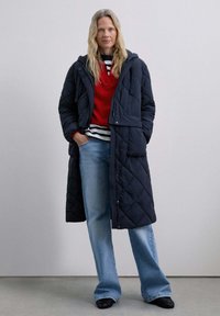 Quilted long navy coat with a hood; layered over a red sweater and striped shirt. Light blue flared jeans and black shoes complete the outfit.