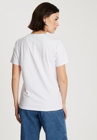 Kobieta z krótkimi ciemnymi włosami, nosząca prostą białą koszulę z krótkim rękawem i niebieskie dżinsy, stojąca bokiem, z jej plecami w większości widocznymi.