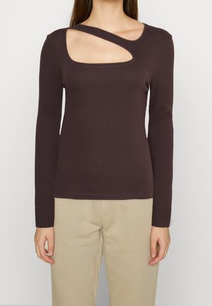 Vrouw die een donkerbruine geribbelde top met lange mouwen en een asymmetrische halsuitsnijding draagt, en beige broek, staand tegen een effen achtergrond.