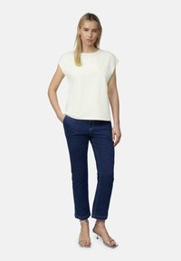 White short-sleeved shirt with embossed detail, paired with dark blue straight-leg jeans. Model stands in silver heeled sandals.