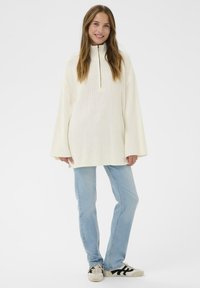 Pull en tricot blanc avec col montant et demi-fermeture éclair, doté de manches larges. Associé à un jean bleu clair et des baskets avec accents noirs.