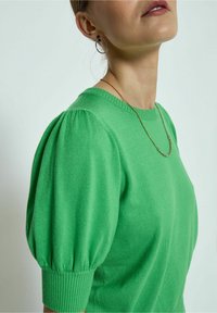Grüner gestrickter Pullover mit kurzen Puffärmeln, einem gerippten Ausschnitt und einer glatten Textur. Akzentuiert mit einer zarten goldenen Halskette.