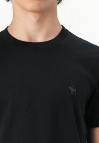T-shirt en coton noir avec col rond, arborant un subtil logo de moose brodé en gris sur le côté gauche de la poitrine.