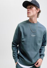 Groene sweatshirt met een ronde hals en logo-tekst op de voorkant, gecombineerd met een zilveren kettinghanger en een zwarte pet. Gladde textuur.