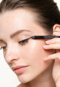 Eyeliner nero tenuto contro la pelle, applicando una linea a goccia. Il primo piano mostra una pelle liscia con leggeri lentiggini, trucco naturale e labbra morbide.