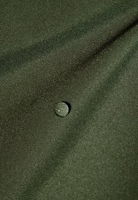 Tissu vert foncé texturé avec un bouton rond recouvert de tissu assorti, cousu au centre créant un effet capitonné.