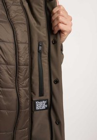 Veste rembourrée marron avec une poche zippée, des fermetures à pression et une étiquette. L'intérieur est matelassé et lisse, mettant en valeur une texture contrastante.