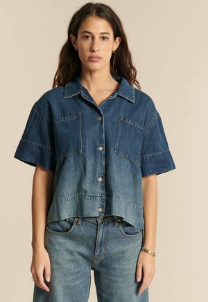 Kobieta o długich ciemnych włosach, ubrana w krótki rękaw koszuli jeansowej zapinanej na guziki oraz dżinsy z wysokim stanem, stoi na tle beżowego tła.