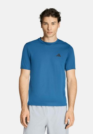 Junger Mann trägt ein blaues kurzärmeliges Sportshirt und hellgraue Shorts und steht vor einem schlichten weißen Hintergrund.