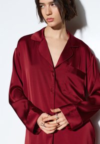 Pudrově vínový saténový pyžamový top s klopovým límcem, jednou náprsní kapsou a knoflíky vpředu. Měkká textura s jemným leskem.