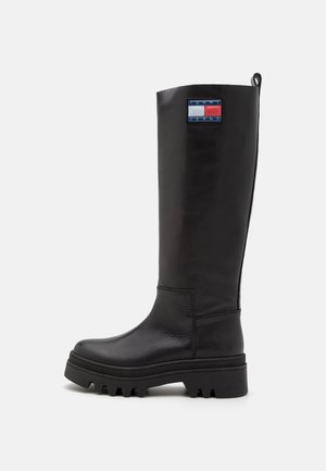 Botte en cuir noire montant jusqu'au genou avec semelle épaisse et patch logo Tommy Jeans près du haut, vue de côté sur un fond clair.