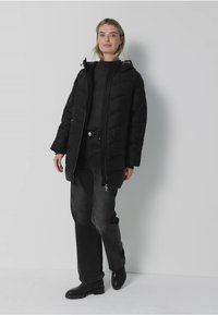 Czarna ocieplana kurtka z zamkiem z przodu, długimi rękawami i wysokim kołnierzem, noszona na ciemnym swetrze, zestawiona z czarnymi dżinsami i botkami.