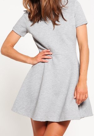 Kvinde iført lys grå, ribstrikket kjole med korte ærmer og knælængde, med en udvidet nederdel, poserer med den ene hånd på hoften og den anden afslappet ned langs siden.