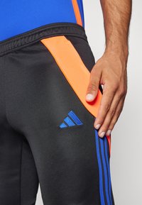 Schwarze Sporthose mit orangefarbener Seitenblende, ausgestattet mit einem blauen Drei-Streifen-Design und einem gestickten blauen Logo auf dem Oberschenkel. Der Stoff wirkt glatt.