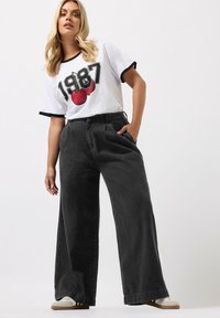 Valkoinen t-paita mustilla reunoilla, jossa on punainen kirsikkakuva ja "1987"-teksti, yhdistettynä löysiin mustiin corduroy-laihoihin housuihin ja beigeihin jalkineisiin.