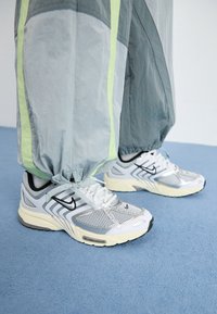 Osoba v šedých teplákových kalhotách s neonovými pruhy a bílých teniskách Nike Air stojící na modrém koberci.